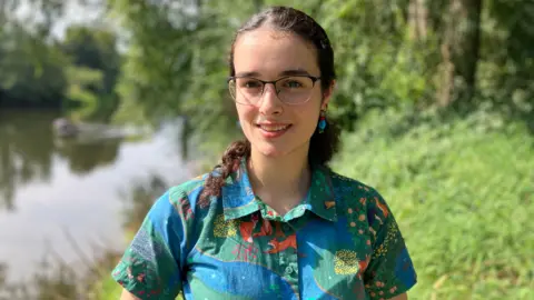 A woman with dark curly hair, wearing glasses and a brightly coloured shirt, stands in front of a river.