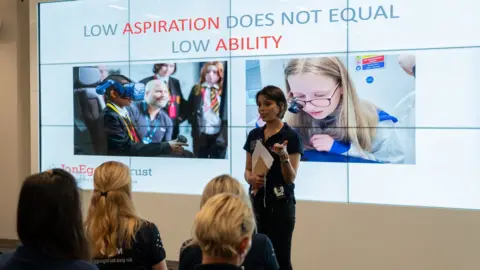 Jon Egging Trust Dr Emma Egging addresses an audience of young people. Behind her, a big screen shows images of children participating in science projects, while a message reads: "Low aspiration does not equal low ability".