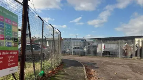 Recycling centre's sign showing what can be recycled at site. It is on metal gates that have cars and vans parked behind it.