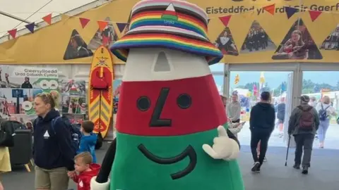 Mister Urdd mascot. He is in the shape of a triangle, with a green base, red middle, and white top. He wears a rainbow bucket hat and stands in a marquee.
