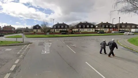 Google A man and woman with white hair and wearing dark trousers and jackets walk across a street junction hand in hand. Houses can be seen on the far side of the road. It is a cloudy day, with small sections of blue sky.