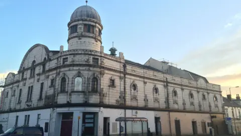 Abbeydale Picture House exterior. It is a fairly magnificent building. The main facade is on a corner, close to a road. Its exterior is decorated with white tiling. It has many windows with curved tops and tiles framing them. There is a taller "tower" section at the front of the building, with a glass dome at its top. It appears run down, and slightly dirty, but clearly was once an impressive building.