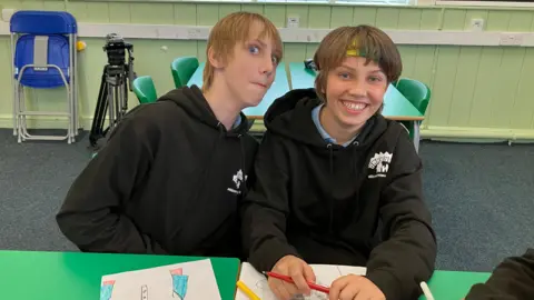 Twin Toady, who has light brown hair, sits at a desk next to Noah, who has slightly darker hair and is wearing a green and yellow headband. They are wearing black school leavers hoodies