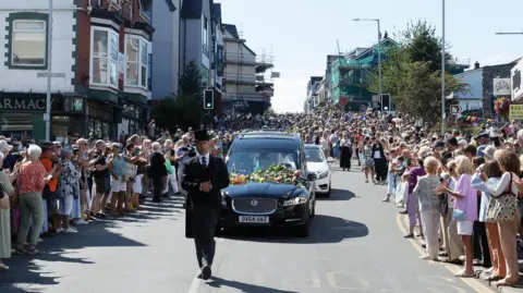 PA Media Thousands of fans gathered in the street as Bonnie Tyler's coffin drives down the road in a black funeral car.