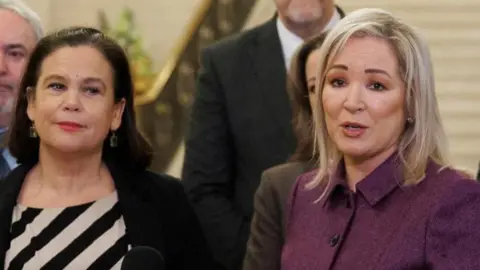 Pa Media Mary Lou McDonald, with brown hair, wearing a white and black top top, standing next to Michel O'Neill, with blond hair, wearing a purple jacket. 