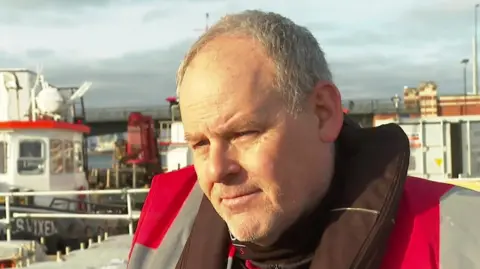 Darryl Fleming is standing in front of barges that will carry the fireworks. He is a man of about 50 with short silver hair and he is wearing a red and grey lightweight waterproof jacket.  