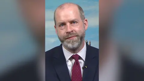 Jonathan Reynolds sat in a studio wearing a suit and a tie