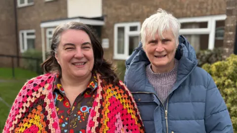 Ben Schofield/BBC Louise and Janet Brown standing side by side and looking directly down the camera. They are in a residential setting, with brick buildings, grass and bushes behind them. They are both smiling. Louise, on the left, is wearing a brightly coloured knitted cardigan, over a flowery patterned top. Janet, who has short grey hair, is wearing a blue winter coat with a hood, with a lilac coloured jumper underneath. 
