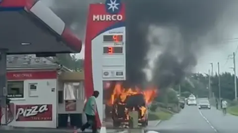 Fin James A car is on fire next to the petrol station, it has dark plumes of smoke coming from it and orange flames. A person is seen looking on. There is a sign that says MURCO