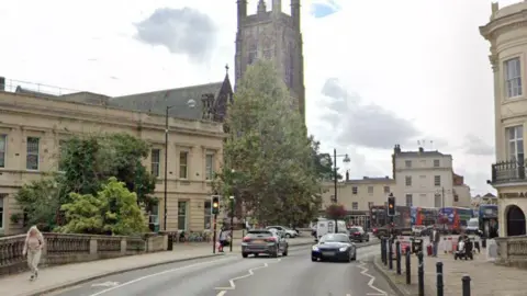 A street with beige-coloured buildings and a church on the left hand side with black bollards lining the left side of the road, cars on the road and people walking on the pavement