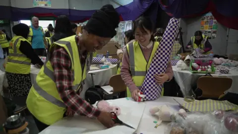 BBC Volunteers wearing high-visibility vests wrap Christmas presents at tables inside a community hall, using scissors and patterned paper, with toys and wrapping materials spread out around them.