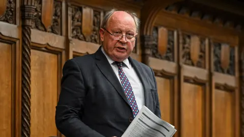 Jim Allister is speaking in the House of Commons.
He is a bald man and wears wire-framed glasses. He is wearing a dark grey suit, pale shirt and a red, white and blue tie. Behind him is wooden panelling.