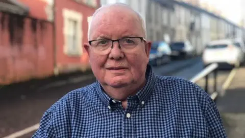 BBC Patrick Gill stands on a street. There are cars and terraced houses behind him. He has grey hair and wears glasses and a blue checked shirt. 