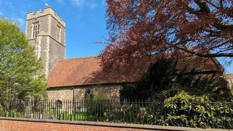 Sandy Gerrard/Geograph A large church with a flint roof and tower. It has graves, bushes and tall trees in its graveyard. There is a brick wall with a black iron fence around the perimeter of the grounds.