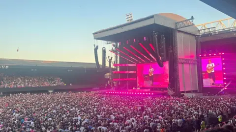 The large stage is placed at the end of the football pitch and is lit up pink. Ed Sheeran stands in the middle of it, wearing a white T-shirt and holding a guitar. The pitch is covered by the crowd, with people also watching on from seats in an adjacent stand.