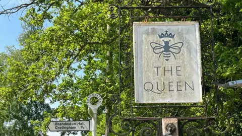 BBC Pub sign "The Queen" with road sign in background