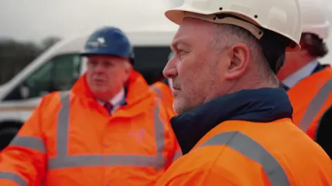 BBC Steve Reed stands to the right of the image, he is looking to the left, slightly away from the camera and has a slight frown. He wears a white protective plastic helmet and orange high vis long sleeved coat, with silver reflective stripes and a navy blue collar, he has grey short hair. The background is slightly blurred, there is a man wearing similar protective gear with a shirt and tie on underneath and a white van in the distance.