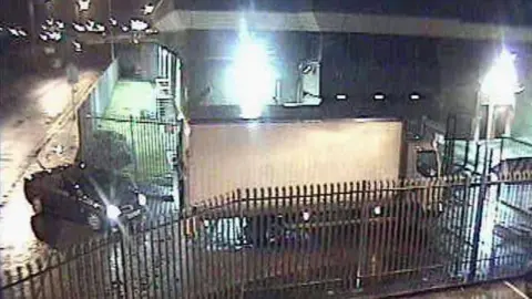Getty Images A handout photo from Kent Police shows a car and a white lorry at the entrance of the Securitas depot in February 2006. There are bright lights outside the depot and the car has its headlights on. The area around the depot is lit up.
