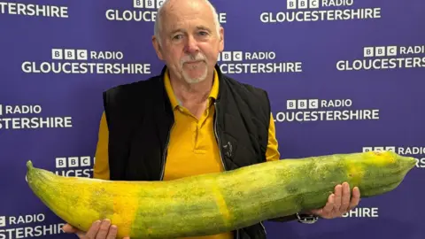 A man carrying a large cucumber in front of the BBC Gloucestershire branding.