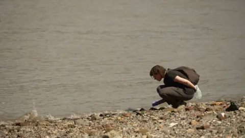 Woman bends down   to inspect thing  connected  enactment     of the Thames