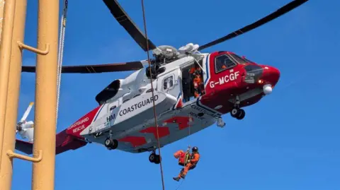 A red and white painted coastguard helicopter performing a rescue operation. The helicopter is hovering mid-air with its side door open, and a rescuer in an orange suit being lowered on a cable toward the ship. The weather appears to be clear and sunny, with a bright blue sky.