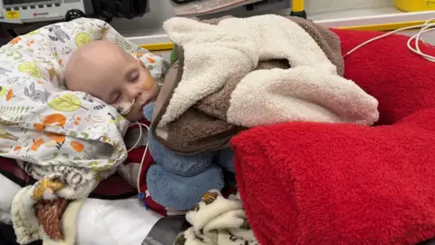 Sarah Sloman A young boy with a feeding tube in his nose lying down with his eyes closed. He has lots of different coloured blankets piled on top of him.