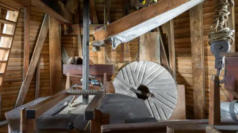 Historic England Archive An octagonal wooden room with the edge of a ladder and wooden planks propped up against wooden slatted walls and different angles, there is a wheel-shaped object on the ground and rope tied around a vertical leaver. The edge of a red sign with white letters is visible, it looks like it reads 'alarm'.