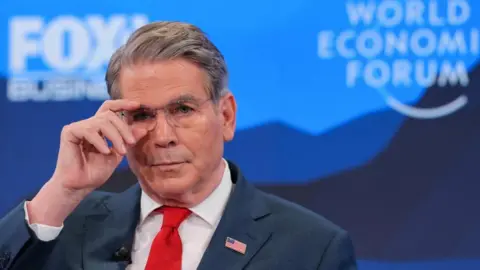 Treasury Secretary Scott Bessent adjusts his glasses on stage at the World Economic Forum. He wears a blue suit and red tie and a flag lapel pin