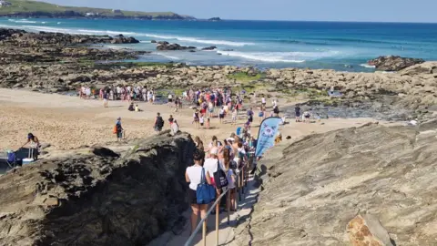 Newquay Beach Care A large crowd gathers on a rocky beach and shoreline, with people queuing down steps towards the sand beside clear blue sea.
