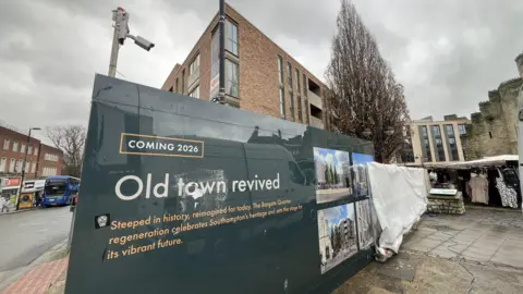 A sign around a newly built apartment block in the centre of Southampton with the bargate visible on the right-hand side. The sign reads "Coming 2026" and "Old town revived... steeped in history, reimagined for today. The bargate quarter regeneration celebrates Southampton's heritage and sets the stage for its vibrant future".