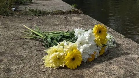 White and yellow flowers laid on a stone by a body of water on a sunny day.