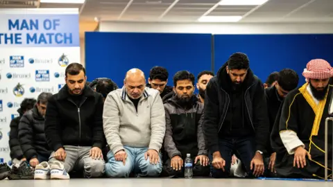 Group of men sat praying. They are all kneeling with their hands on their knees and their heads bowed.