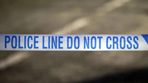 A close up of police tape that reads "Police line do not cross". The road is blurred in the background.