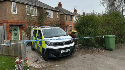 A police van is parked on the drive of a house. Police tape stretches across the drive from a green bin to a fence panel. Some flowers have been laid to the left of the drive, by the fence panel. The front door of the house is green with red brick around the lower level of the house it and brown render above.