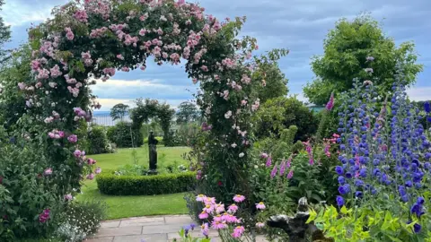 David Hines/Liz Kelly-Hines Large garden with lots of bright pink, purple and blue flowers in the foreground. The flowers are of all different varieties and there is a very neat lawn with a statue in the middle of it in the distance - behind an archway of pink roses.