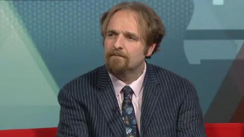 Robert Alden is the councillor for Erdington and leads the Birmingham Conservatives. He is wearing a stripy jacket, pink shirt and a blue, funky tie. He has medium-length hair and a goatee.