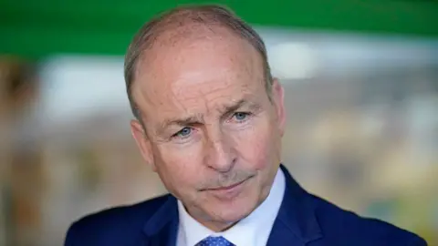 PA Media Micheál  Martin with balding light brown hair and blue eyes wearing a dark navy suit and looking towards someone speaking to him