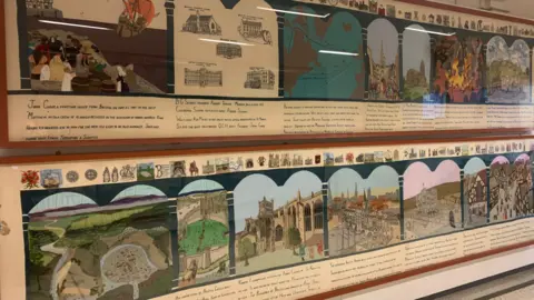 A wide shot of the framed Bristol Tapestry, showing its long rectangular horizontal panels. There are two panels, one above the each, with each depicting around seven pictures of key milestones in the city's history.