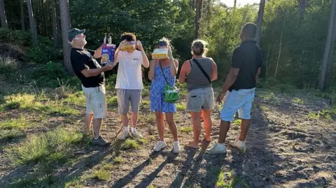 Bob Courtice A group of people stand in a sunny clearing on Ockham Common in Surrey, holding eclipse viewers and projection cards while looking towards the sky. Tall trees surround the clearing and long shadows stretch across the ground.