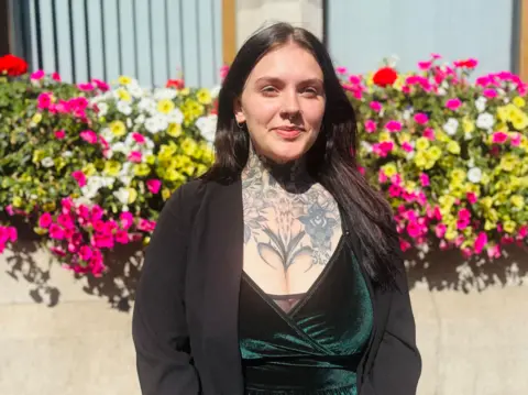 A young woman with dark hair smiling at the camera. She has a large tattoo on her chest and is wearing black clothes. She is standing in front of colourful flowers. 