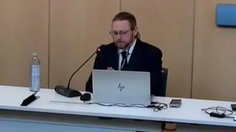 Cambridgeshire County Council Livestream image of James Sidlow. He has long hair tied back, is wearing glasses, has a beard and is wearing a dark suit and tie. He is sitting behind a laptop and is talking into a microphone