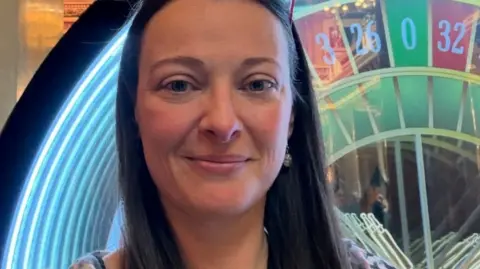 Rhona Paton A woman with long brown hair and sunglasses, she is standing in front on a roulette wheel.