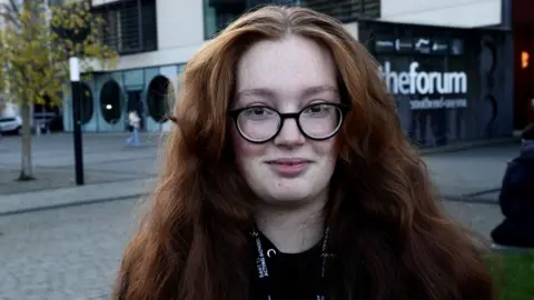 Stuart Woodward/BBC A woman wearing round framed glasses and long brown hair. She is standing at the Southend-on-Sea campus and smiling at the camera