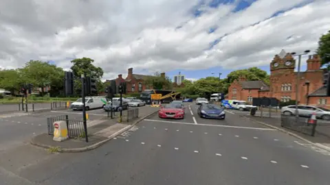 Cars queueing across five lanes at traffic lights, red brick buildings and tress in the background. 