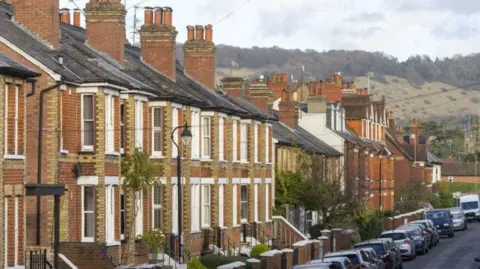 A row of redbrick terraced housing. There is countryside in the background and cars are parked along the street. 