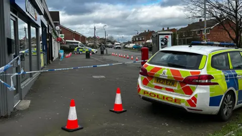 BBC A police tape cordon around a small parking area outside the front of a row of shops. The back of a police car can be seen. Two traffic cones sit behind the police car. Houses are on the other side of the street.