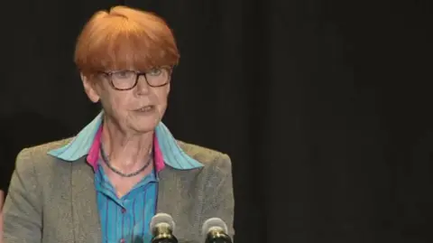 BBC Dame Vera Baird, with short red hair and glasses, is pictured speaking at a press conference wearing a blue shirt and gray blazer.