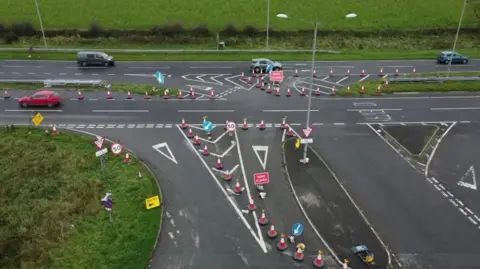 The A30 at Plusha. An aerial shot depicts a traffic management setup at a road junction. The area has many traffic cones, temporary road signs and directional arrows. Speed limit signs for 50mph are in place, as well as diversion arrows directing traffic through a controlled path. The surrounding area has green grass and a rural backdrop.