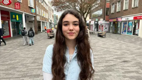 A young woman with long brown hair. She is wearing a pale blue shirt.