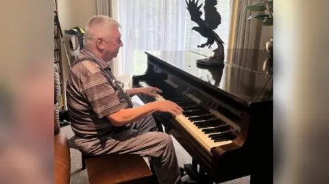 Blind Veterans UK Graham Grange is sat at the piano, with his hands resting on the keys as if he is playing it or about to. There is what appears to be a wooden model of an eagle on top of the piano